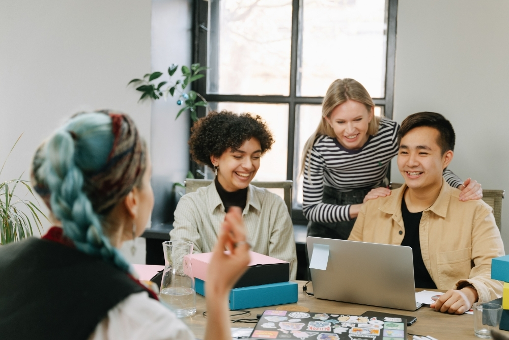 Team members smiling during an office meeting, collaborating around a laptop at a desk.